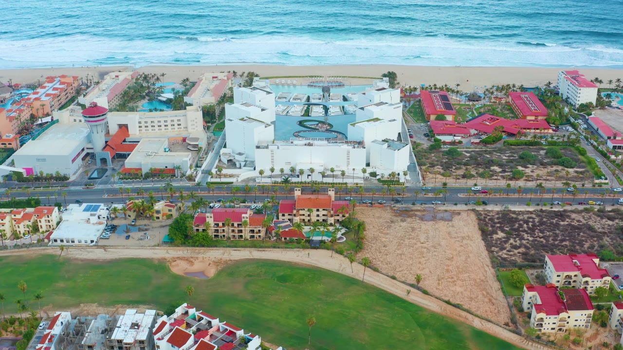 Wide aerial of coastal hotel buildings, roadway, beach, ocean, and lawns.