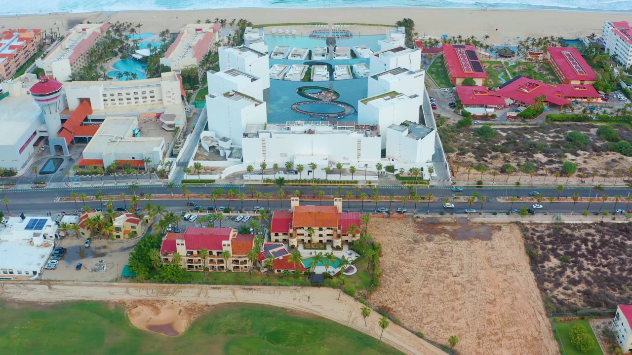 Aerial frame in San Jose del Cabo with breaking ocean waves at the shoreline, a white beachfront complex, red-roofed buildings, a parallel road with small vehicles, and no visible people.