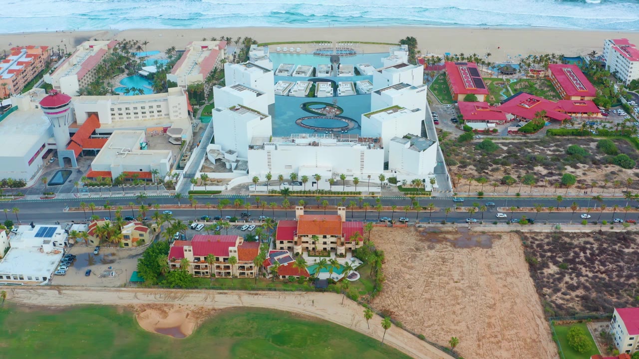 Aerial view of beach surf, beachfront hotels or residences, coastal road, and grass.