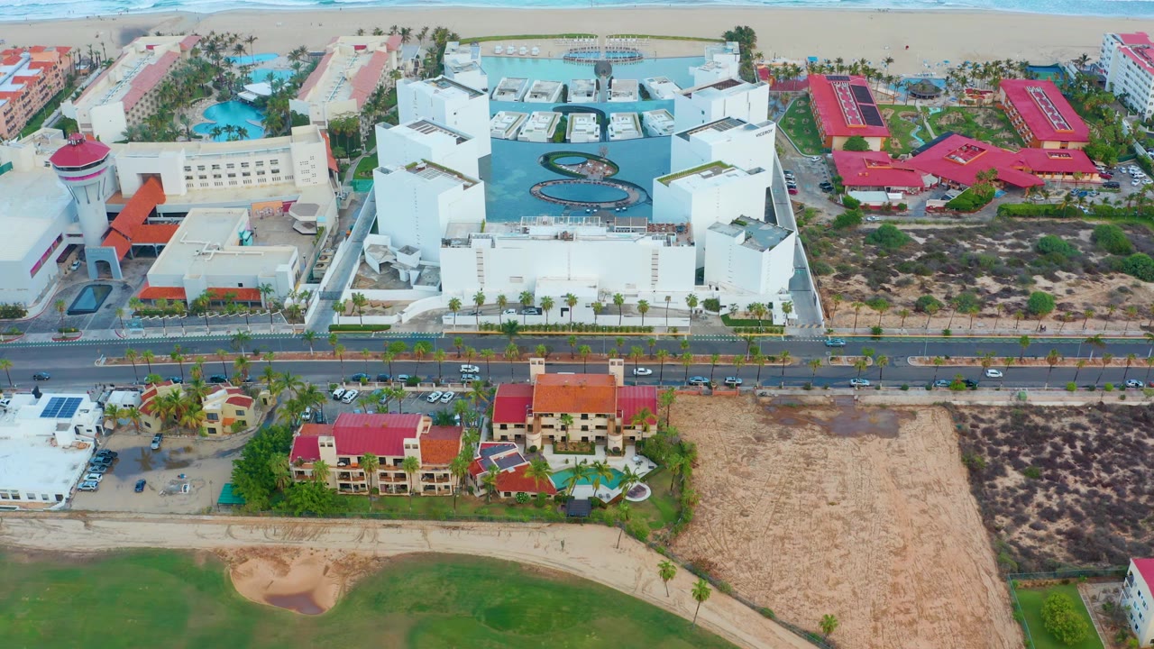 Opening aerial frame over San Jose del Cabo coastal buildings, with ocean surf along the top edge, white pool buildings near center, red roofs nearby, a road across the scene, and no visible people.