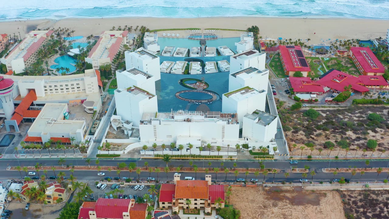 Opening aerial frame over San Jose Del Cabo showing white resort-style buildings, blue pools, a road with cars, red-roofed structures, sandy beach, pale ocean water, and no visible people.