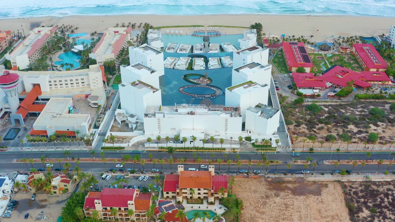 San Jose Del Cabo aerial frame showing the coastal road across the lower third, white resort buildings and pools near center, red-roofed buildings on both sides, sandy beach and ocean behind them, and no visible people.