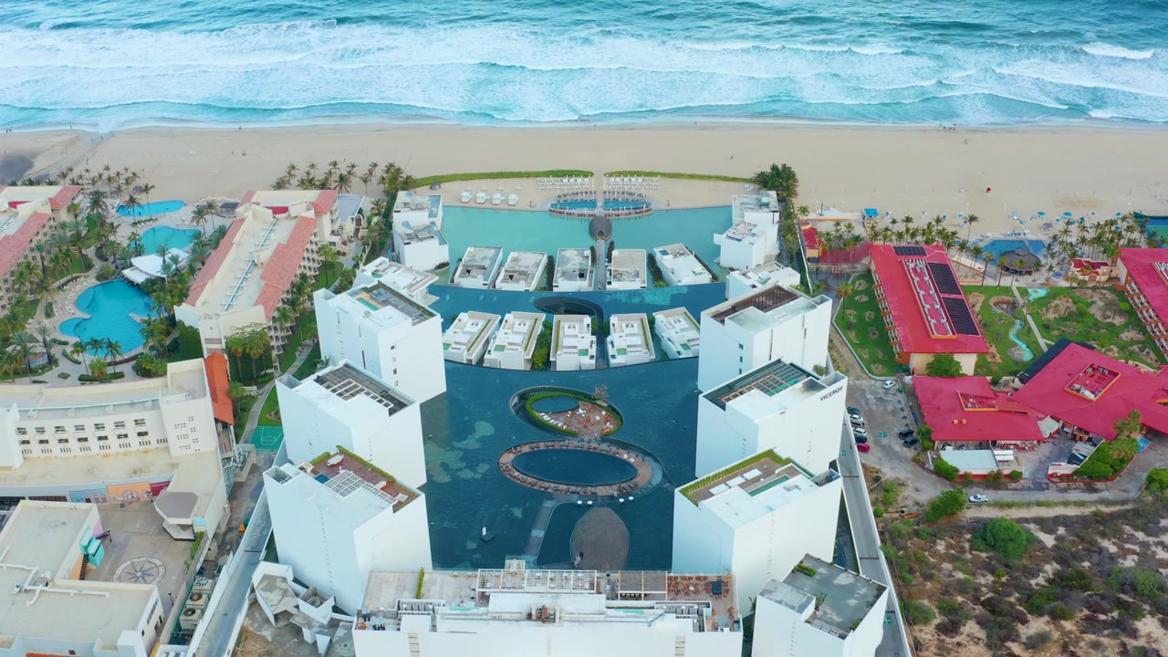 Aerial frame over a San Jose del Cabo resort with white buildings around dark blue pools, red-roofed structures nearby, sandy beach and surf beyond, and no visible people.