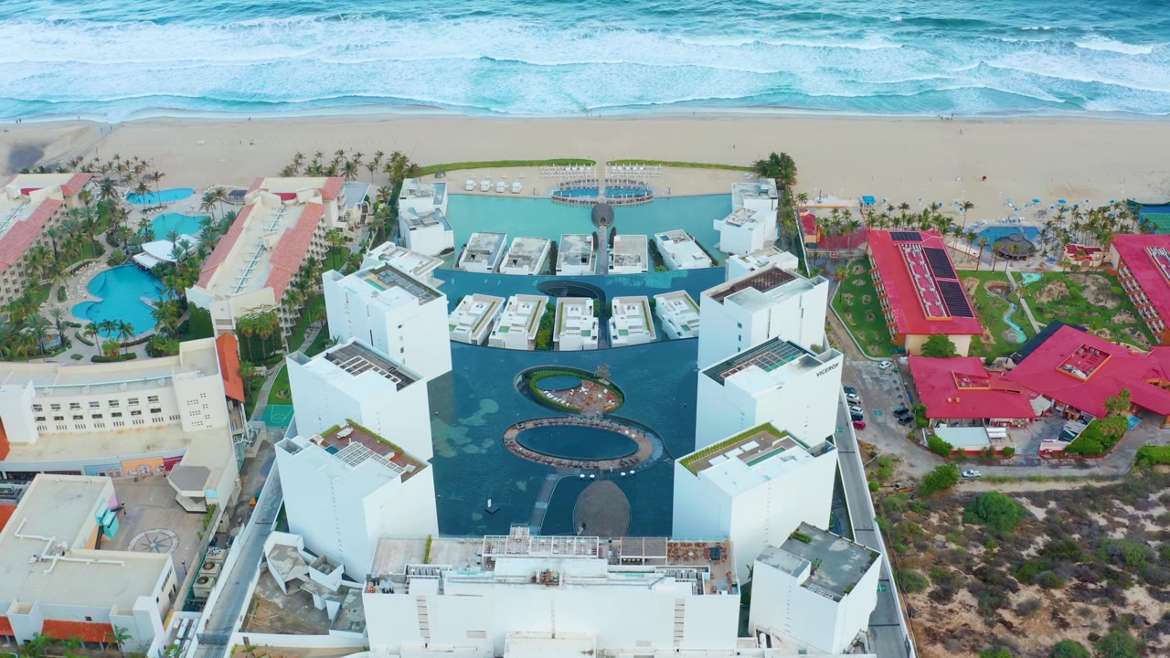 San Jose del Cabo aerial frame showing the white resort angled between a blue pool area and red-roofed neighboring buildings, with beach and surf behind and no visible people.