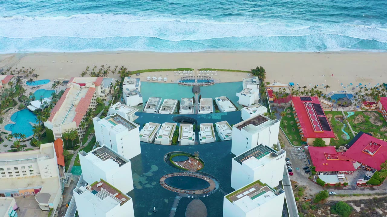 Opening aerial frame in San Jose Del Cabo showing white resort buildings around blue pools below a sandy beach and bands of turquoise surf, with a tiny person visible on the shore.