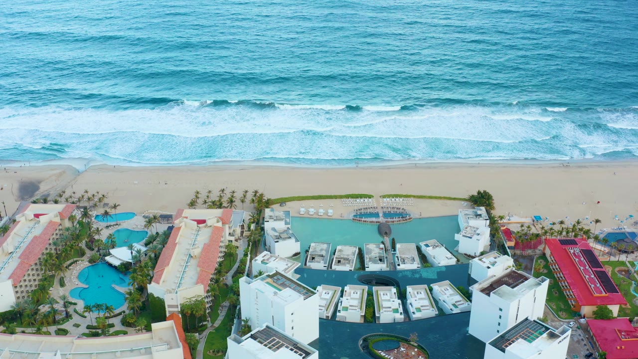 Aerial view of ocean surf, sandy beach, resort pools, white buildings, and red roofs.