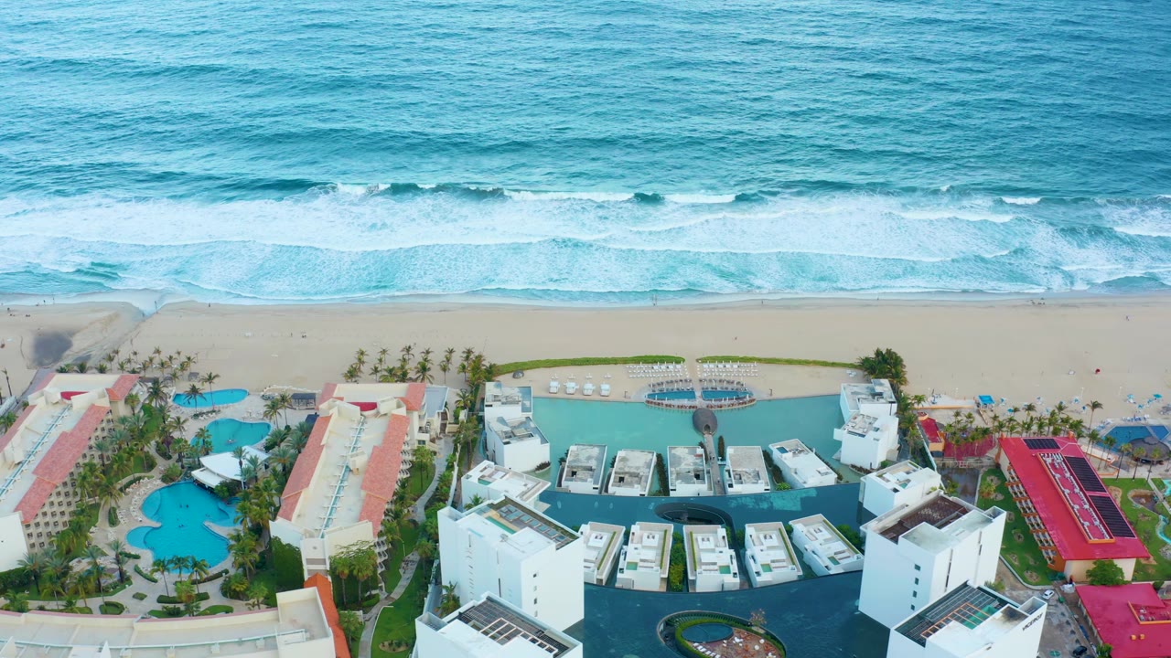 Aerial frame of the San Jose del Cabo resort with parallel surf bands across the ocean, pale sand below them, and clustered pool decks and white buildings in the foreground, with no visible people.