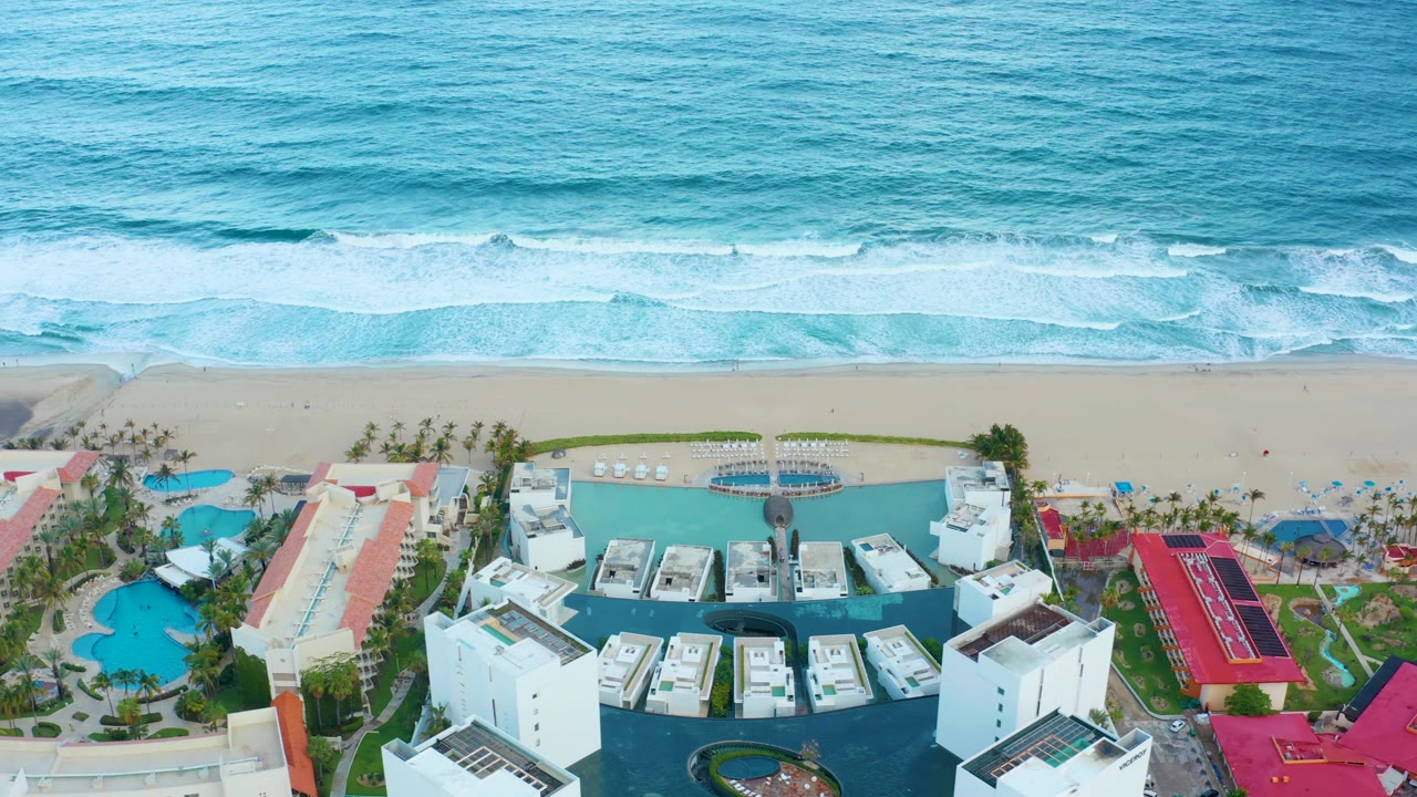 Aerial frame of the San Jose del Cabo shoreline with brighter surf bands, a clean sand strip, and resort pools, palms, white towers, and red-roofed sections below, with no visible people.
