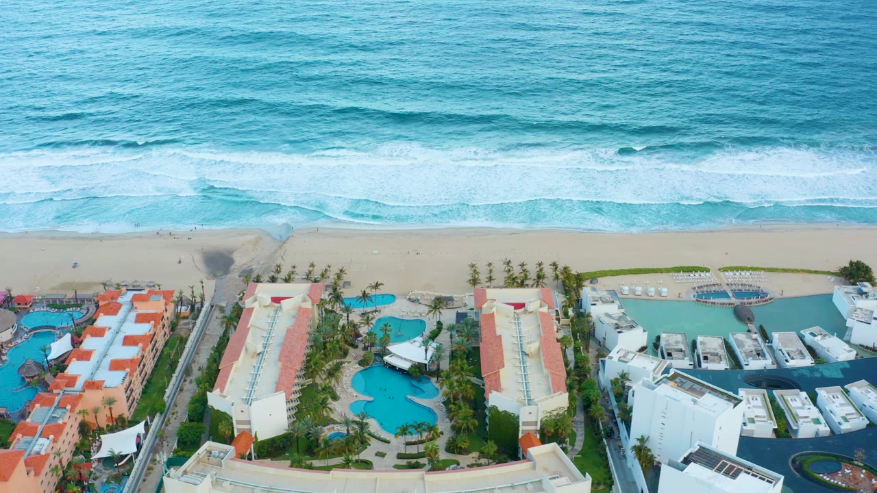 Aerial frame over a San Jose Del Cabo coastal resort, with blue ocean surf above the sandy beach and pools, red roofs, palms, and white buildings below, no visible people.