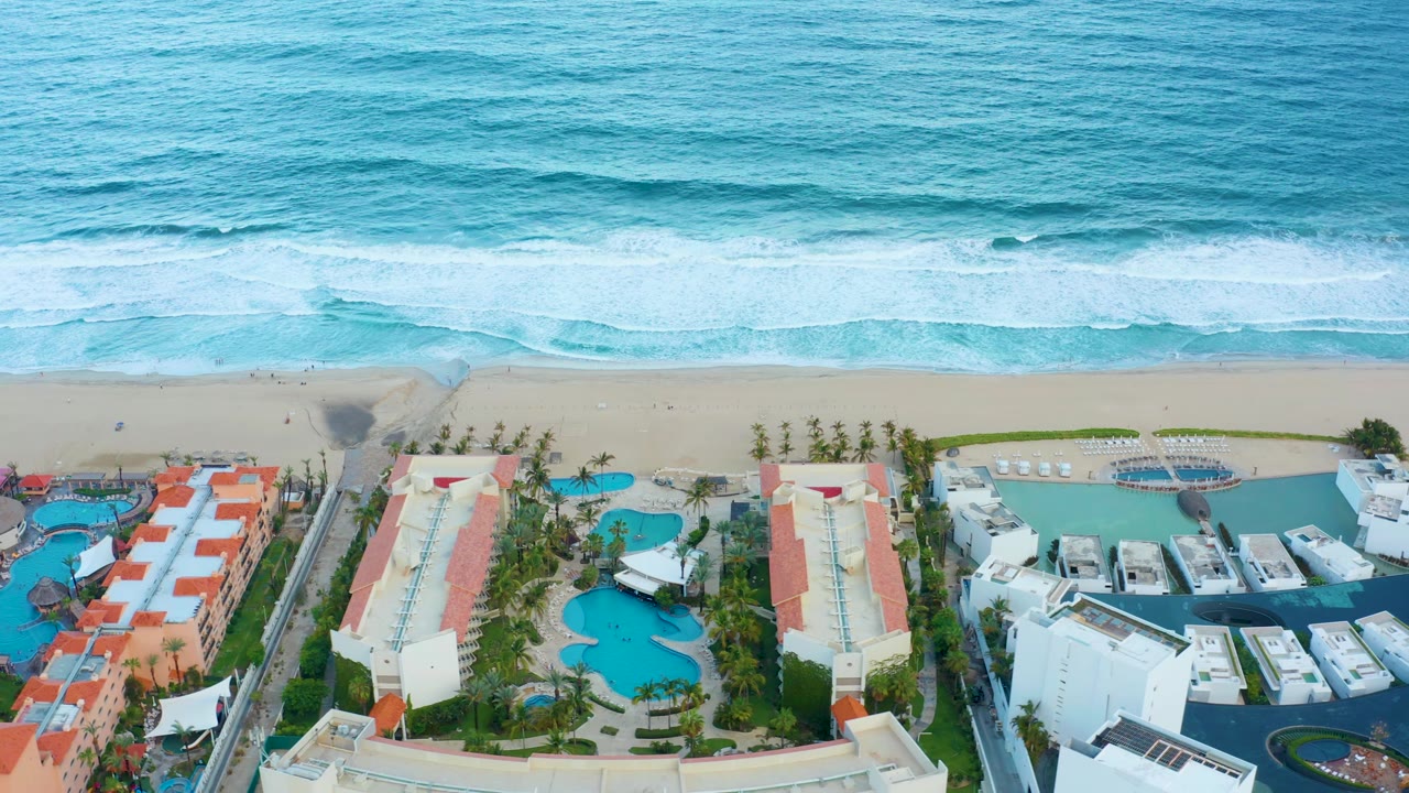 Early aerial frame in San Jose Del Cabo showing a broad blue surf band, pale sand, palm-lined resort grounds, red-roofed buildings, and white hotel structures, no visible people.