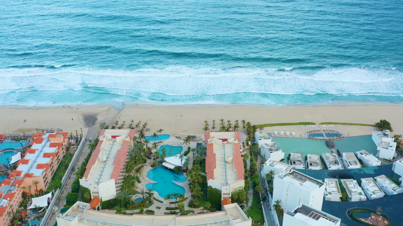Aerial view over the San Jose Del Cabo shoreline as waves line the upper frame and the resort's turquoise pools, white buildings, red-roofed blocks, and palms fill the lower frame, no visible people.
