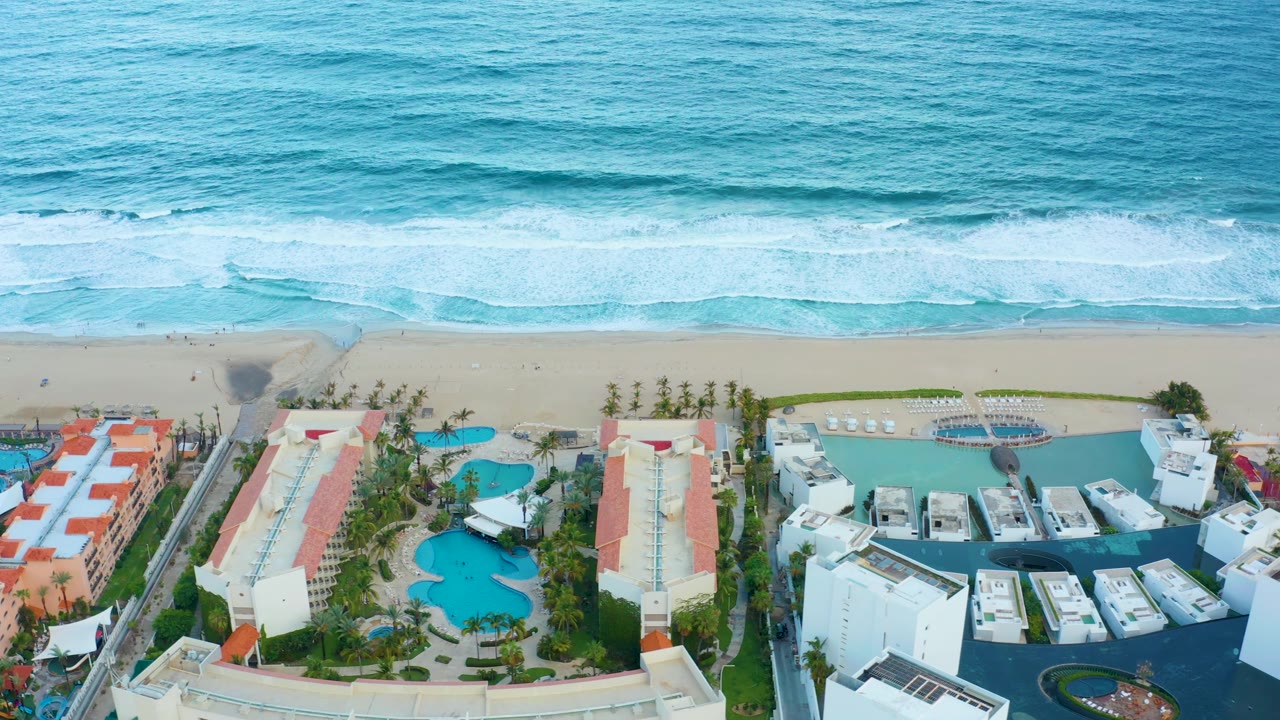 Coastal resort aerial with white hotel blocks, swimming pools, palm trees, sand, and surf.