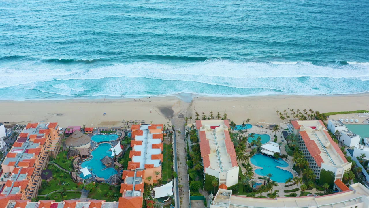 Aerial frame from San Jose del Cabo showing turquoise ocean surf, sandy beach, red-roofed resort buildings, blue pools, palms, and no visible people.