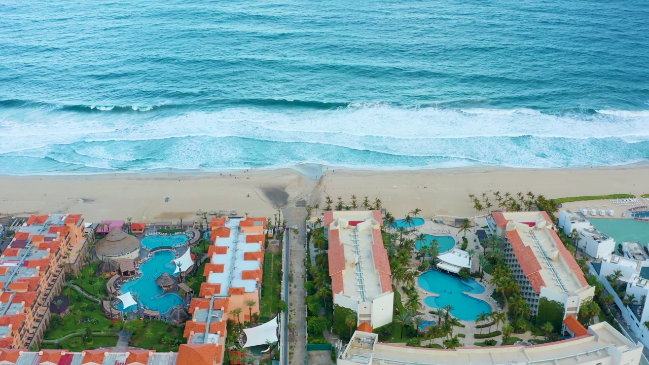 Aerial San Jose del Cabo resort frame showing surf lines across the top, beach in the middle, and white buildings, red roofs, pools, and palms below, with no visible people.