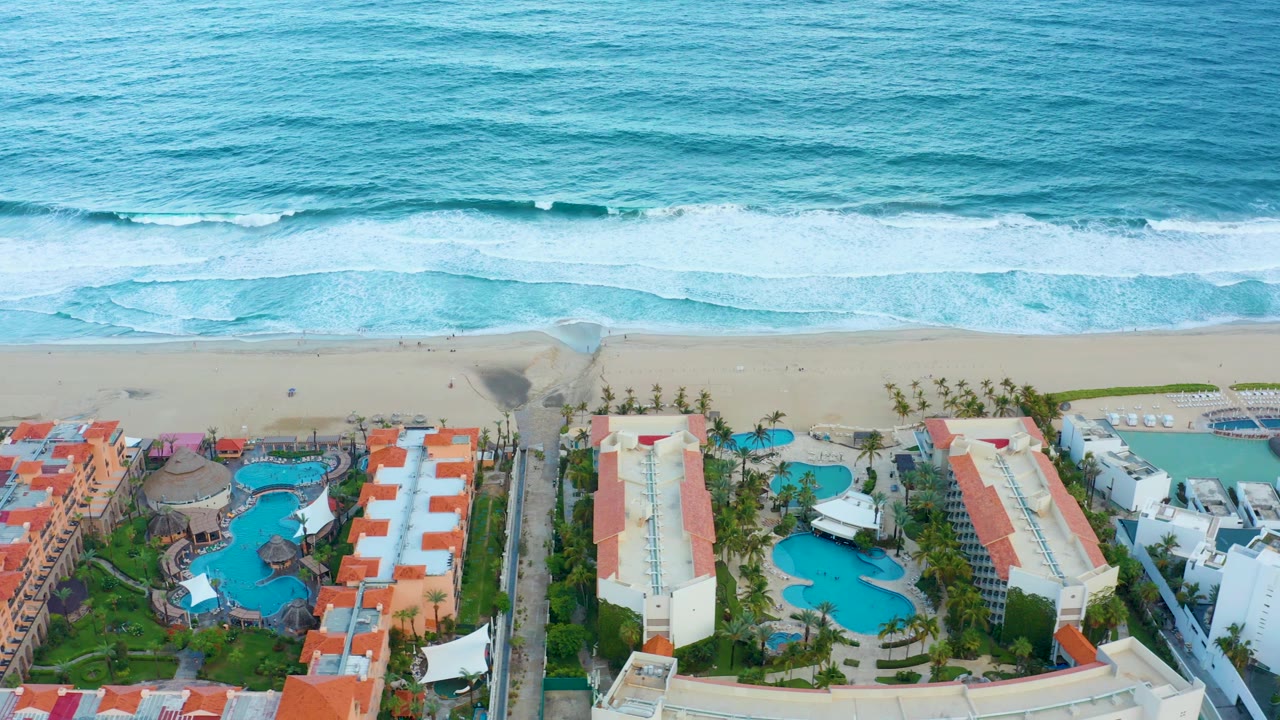 Ocean surf above a resort with blue pools and red roofs.