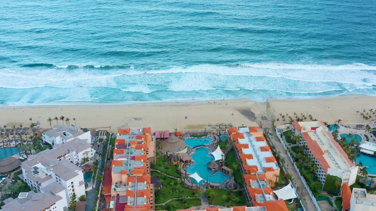 Aerial view of the San Jose Del Cabo shoreline as whitewater rolls toward the sand above resort pools, orange roofs, palm rows, and no visible people.