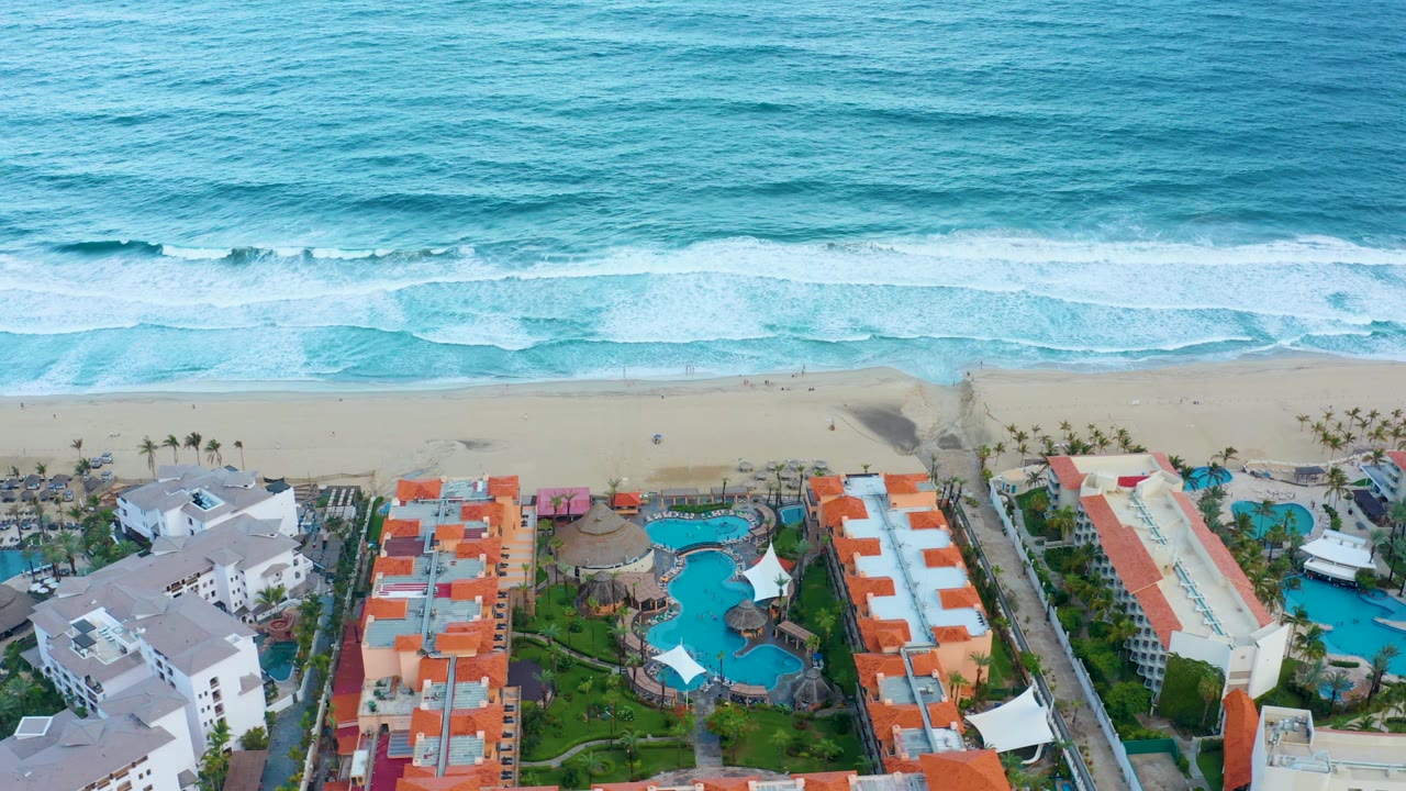 Overhead San Jose Del Cabo beach frame with broad blue water, a bright band of foam, empty-looking sand, resort pools, orange rooftops, and no visible people.