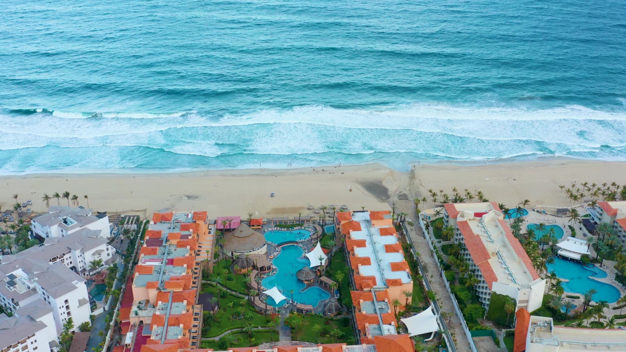 San Jose Del Cabo aerial frame showing ocean texture, white waves breaking near shore, resort blocks, pool water, palms, paths, and no visible people.