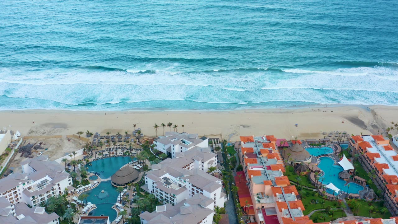 Aerial frame in San Jose del Cabo showing orange-red roof rows beside blue pools, white resort buildings, sandy beach, and layered surf, with no visible people.