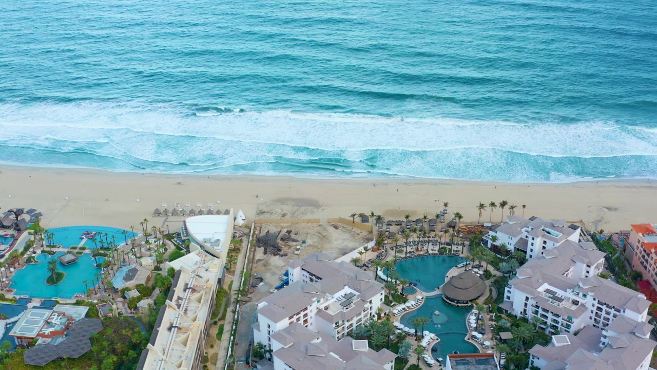 Aerial frame in San Jose del Cabo with foamy waves crossing the top, pale sand through the center, resort pools and white roofs below, and no visible people.