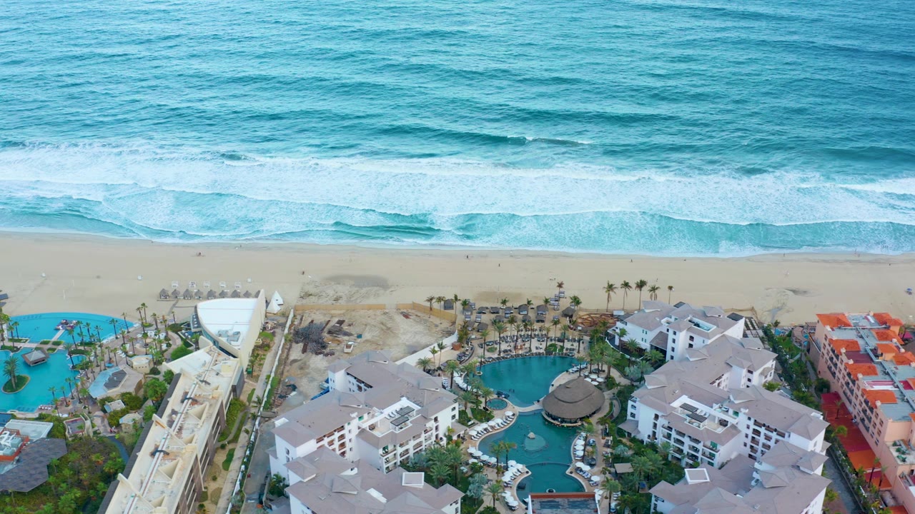 Mid-clip aerial view in San Jose del Cabo where red-roofed buildings become more visible beside blue pools, with ocean surf and an empty-looking beach above and no visible people.