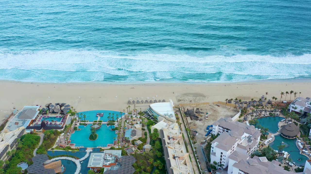 Aerial frame over San Jose del Cabo showing blue ocean waves approaching a sandy beach above resort pools and white buildings, with no visible people.