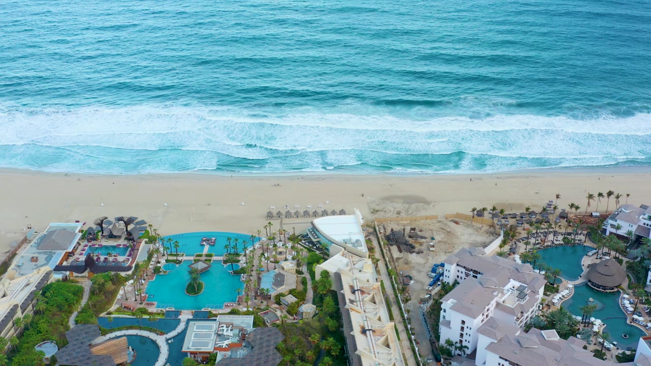 Aerial frame over San Jose del Cabo with a broad band of ocean surf, pale sand, turquoise pools, white resort buildings, and no visible people.