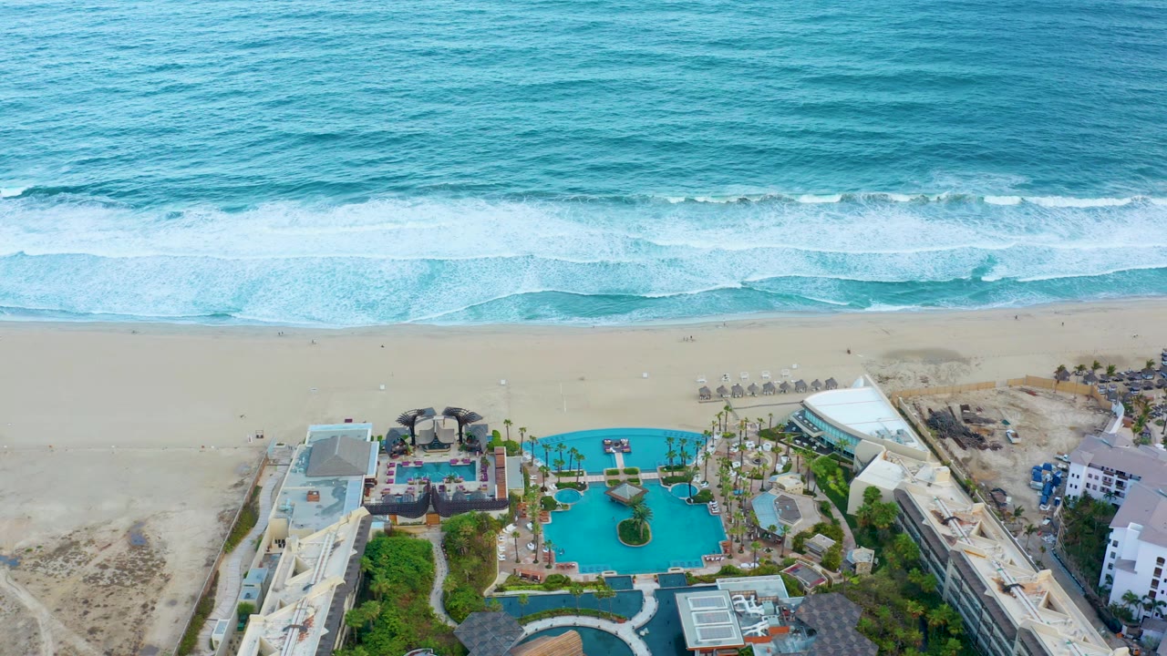 Aerial view in San Jose Del Cabo, Baja California Sur showing blue ocean waves above a sandy beach and a resort pool complex below, with no visible people.
