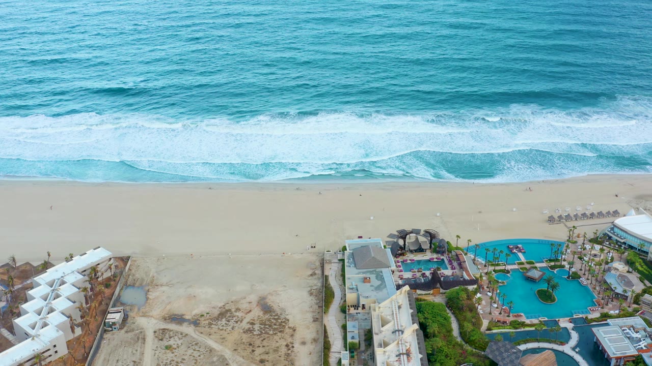 Aerial view in San Jose del Cabo with a wide band of surf, pale sand, and a resort pool area partly framed by white buildings, with no visible people.