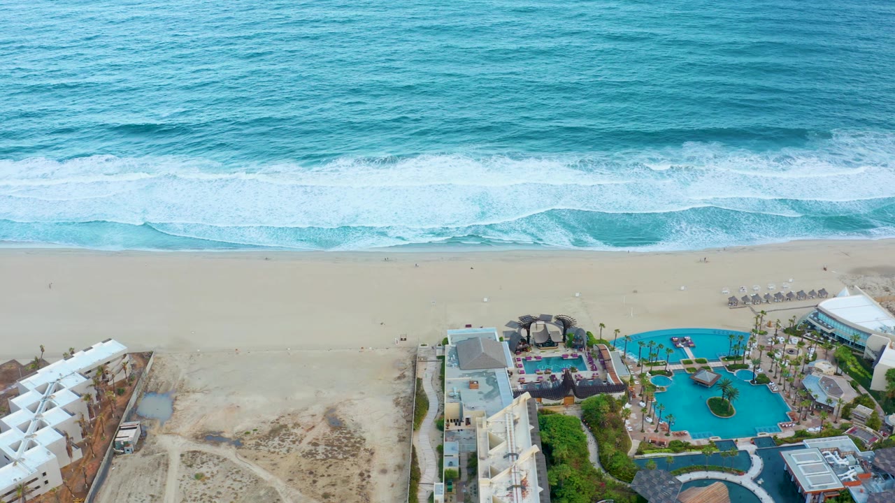 San Jose del Cabo aerial frame showing foamy waves across the top, a mostly empty sandy beach, and blue resort pools bordered by buildings and landscaped paths, with no visible people.