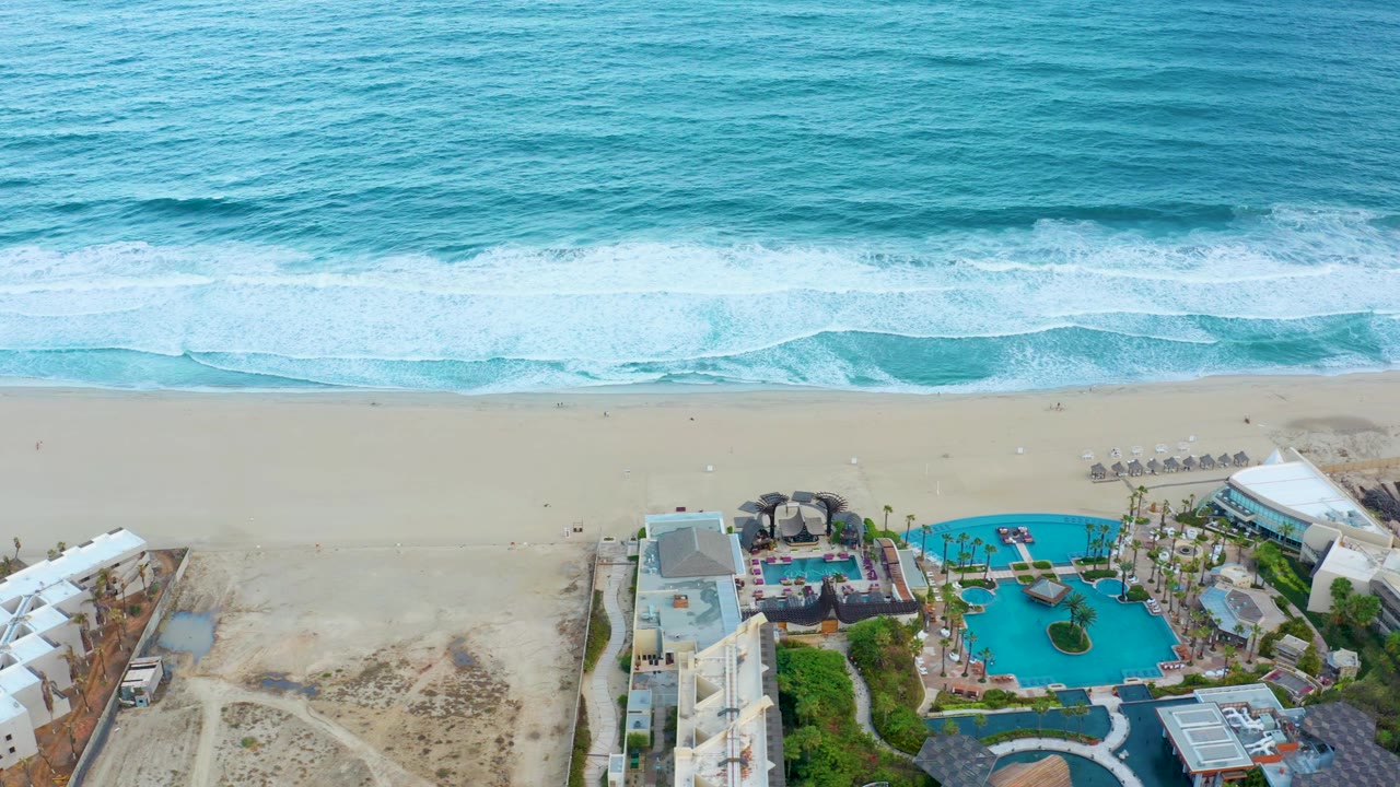 Aerial frame over San Jose del Cabo with rolling blue-green water, a white surf break, pale beach, and resort pools and buildings along the lower edge, with no visible people.