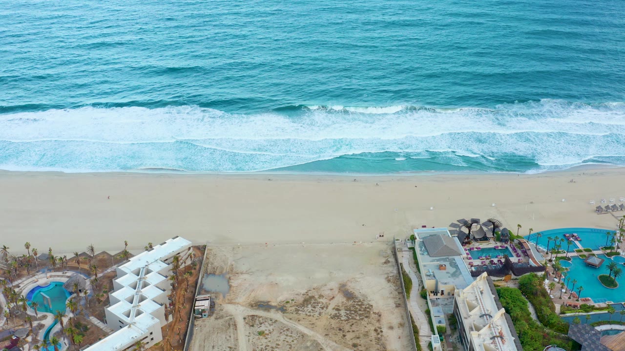 Aerial view in San Jose Del Cabo, Mexico, showing turquoise ocean, white surf, wide sand, resort buildings, blue pools, and no visible people at the start of the clip.