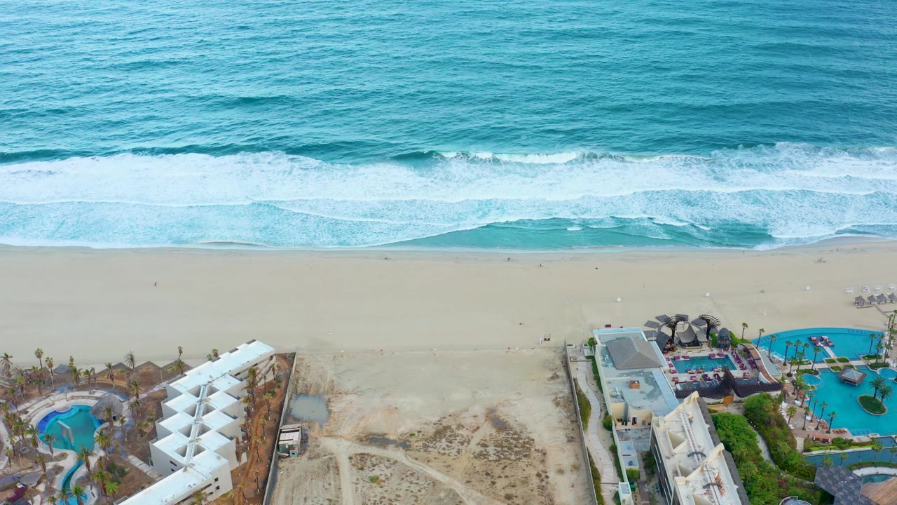 San Jose Del Cabo aerial frame with the surf band shifted along the beach, pale sand below the waves, resort roofs and pools at the lower edge, and no visible people.