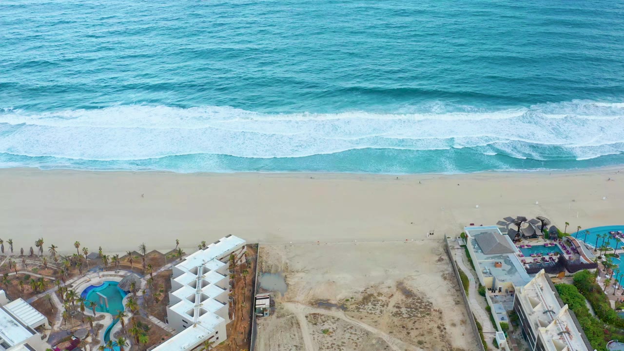 Overhead view in San Jose del Cabo showing turquoise ocean, white surf, pale sand, resort buildings, and blue pools along the lower edge with no visible people.