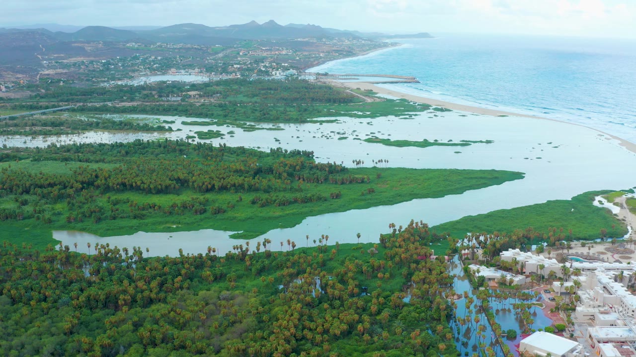 Opening aerial frame over San Jose Del Cabo shows green wetland vegetation and pale river water leading toward a sandy beach and blue ocean, with distant hills and no visible people.