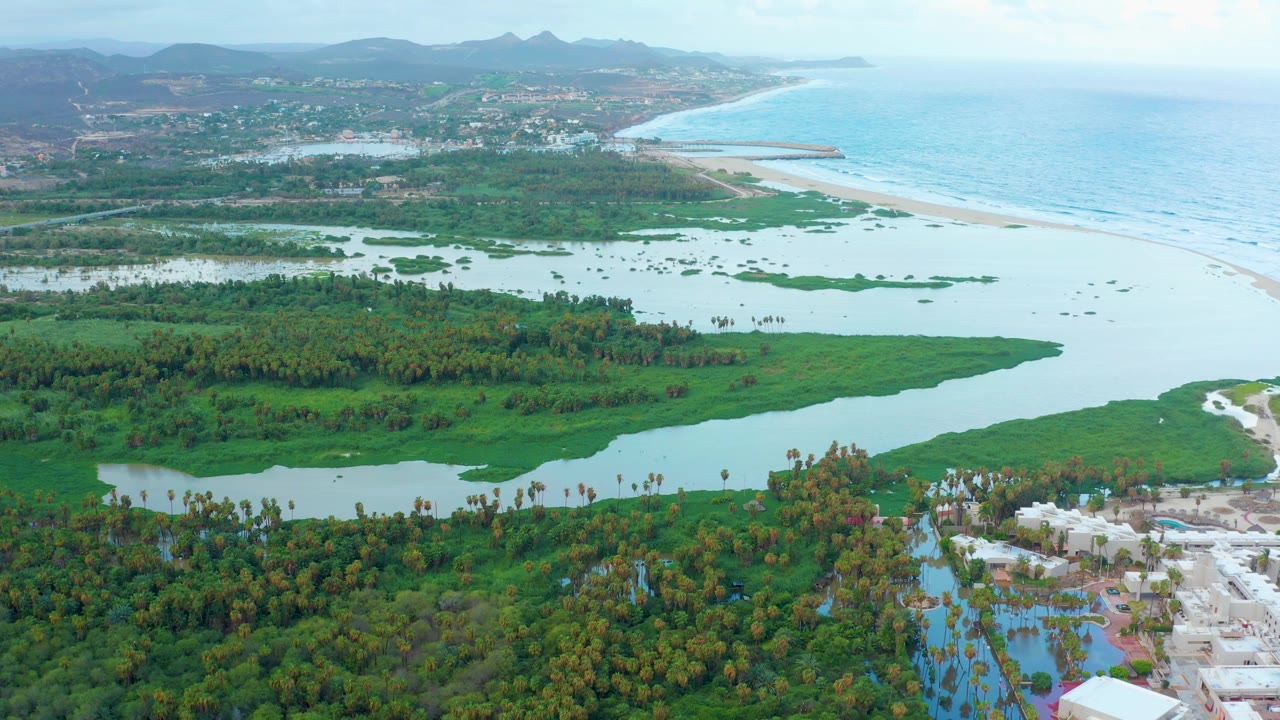 At 0:02, San Jose Del Cabo wetland channels and green islands fill the middle of the frame while the sandy beach and ocean sit to the right, with hills in the background and no visible people.