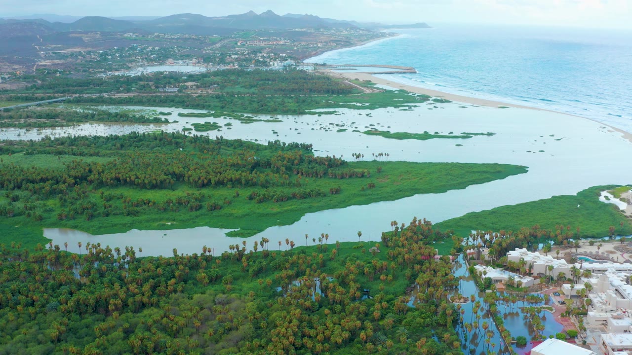 Wide drone view of river water crossing green vegetation toward a pale beach and blue ocean.