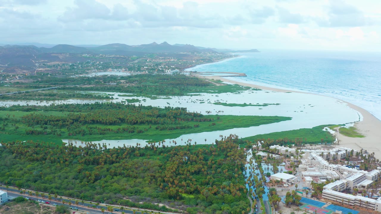 San Jose Del Cabo aerial frame with the lagoon channel running across dense greenery toward the beach and ocean, with distant hills under clouds and no visible people.
