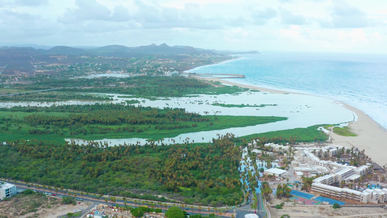 Drone-height view of San Jose Del Cabo, Baja California Sur with the lagoon spread across green vegetation, the beach and ocean on the right, developed buildings at the lower edge, and no visible people.