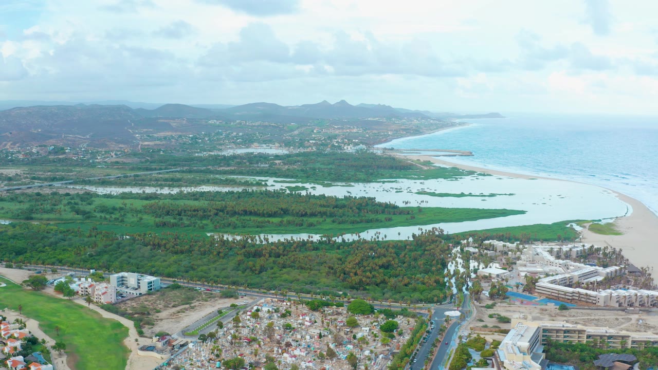 Aerial view over San Jose Del Cabo with resort construction and roads in the foreground, green wetlands and beach in the middle distance, ocean and mountains beyond, and no visible people.