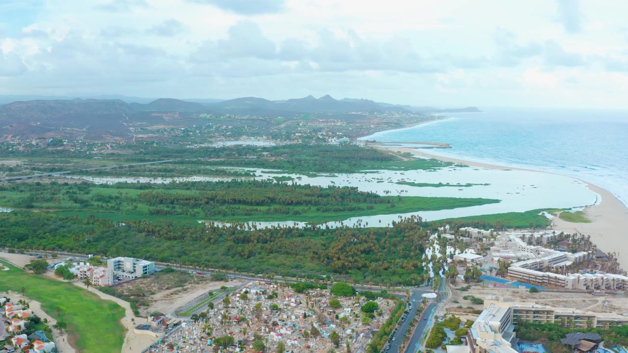 Aerial view over San Jose Del Cabo with the resort construction site near the lower right edge, green wetlands spanning the middle of frame, pale sand and blue ocean to the right, and no visible people.
