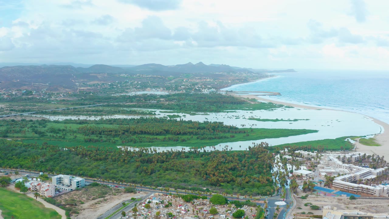 Aerial view in San Jose del Cabo with lagoon water cutting through green land, the beach bending toward the right edge, ocean surf beyond it, foreground buildings, hazy hills, and no visible people.