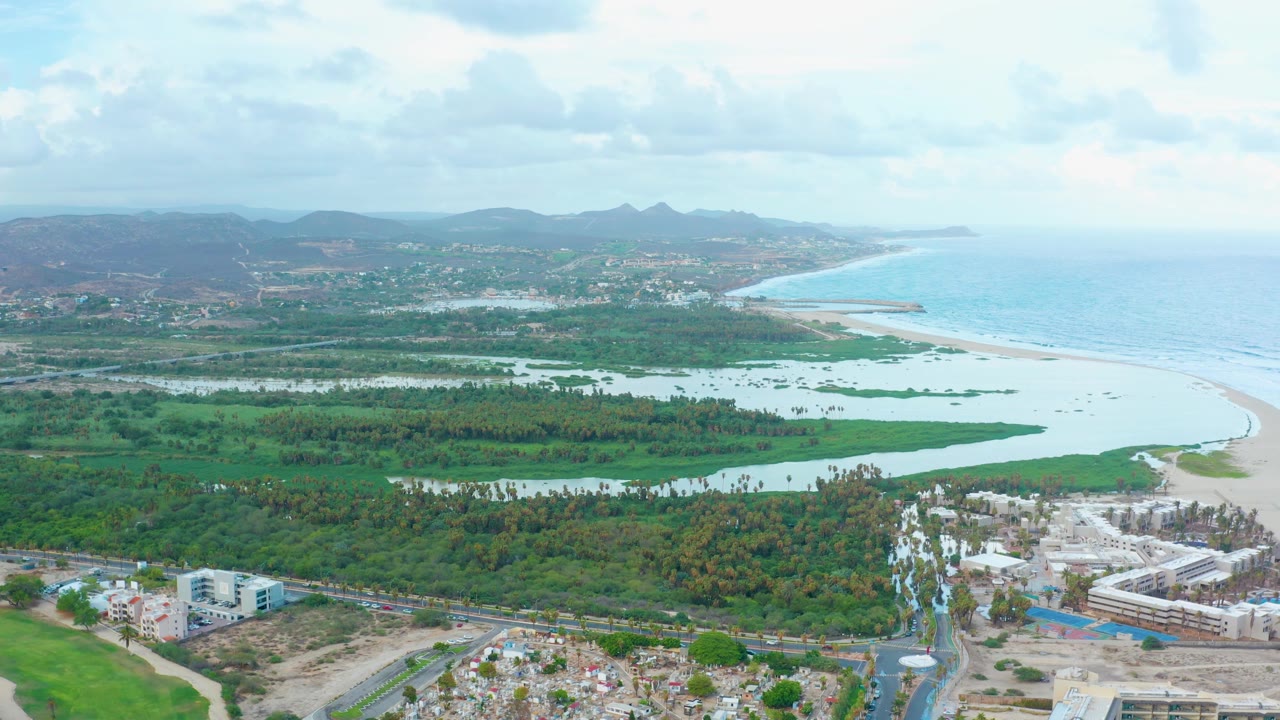 San Jose del Cabo aerial frame showing flooded lagoon areas across dense greenery, a pale sand shoreline beside blue ocean, low-rise construction and roads below, mountains on the horizon, and no visible people.
