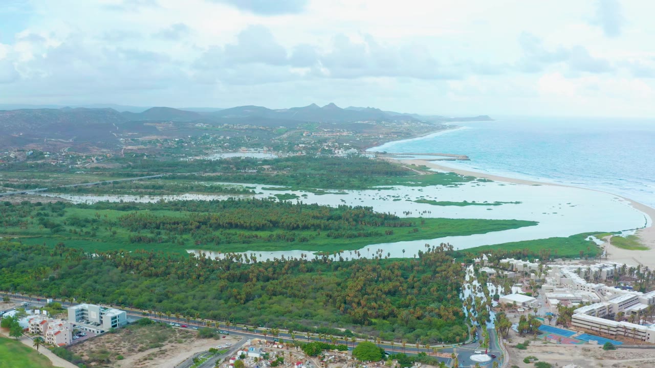 Opening aerial frame over San Jose Del Cabo showing green wetlands around a coastal lagoon, sandy beach, blue ocean, distant hills, road, buildings, and no visible people.