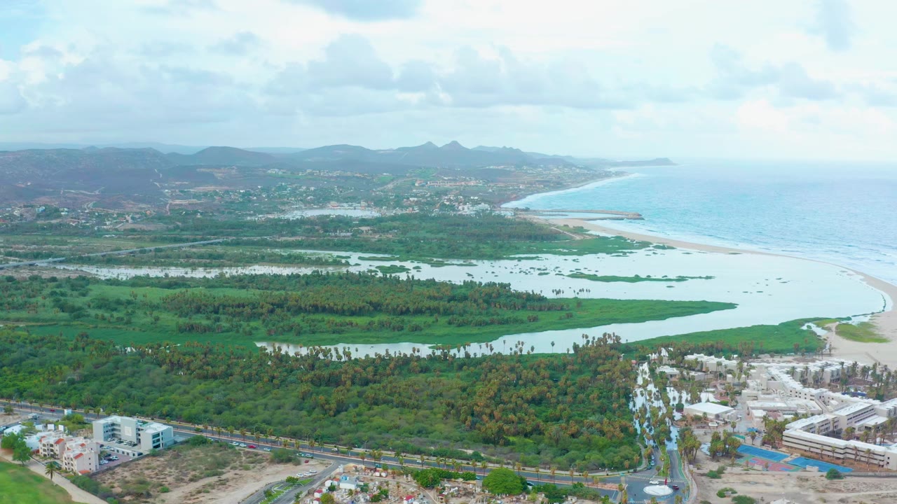 Aerial view in San Jose Del Cabo with the lagoon and green vegetation across the foreground, beach curving toward the ocean, distant hills, buildings, and no visible people.