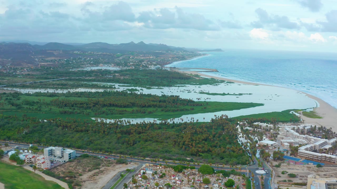 Opening aerial frame over San Jose del Cabo with a pale lagoon crossing green vegetation, a sandy beach and blue ocean beyond, hills in the distance, roads and buildings below, and no visible people.