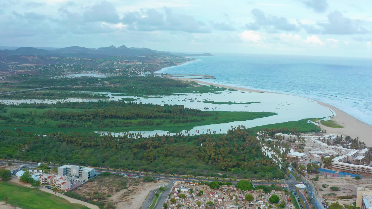 Aerial view over San Jose del Cabo with a pale ocean and curved beach to the right, a reflective lagoon across the middle, low buildings and roads below, distant hills, and no visible people.