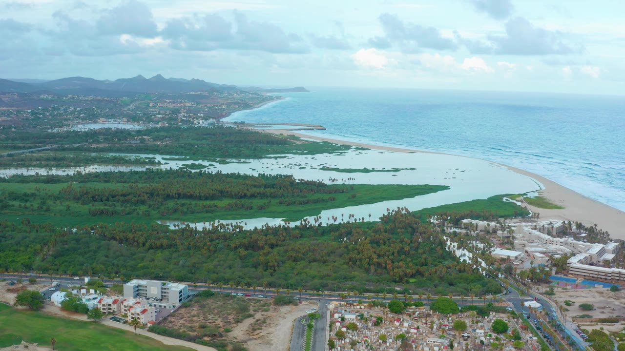 Opening aerial frame over San Jose del Cabo showing a green lagoon beside a curved sandy beach, blue ocean, foreground roads and buildings, distant hills, clouded sky, and no visible people.