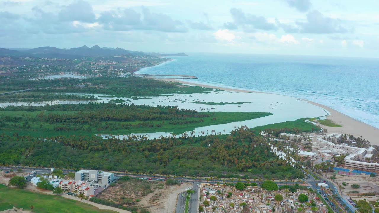 Early aerial frame over San Jose del Cabo with lagoon channels and green vegetation in the midground, a pale sand beach curving along the ocean, foreground development, and no visible people.