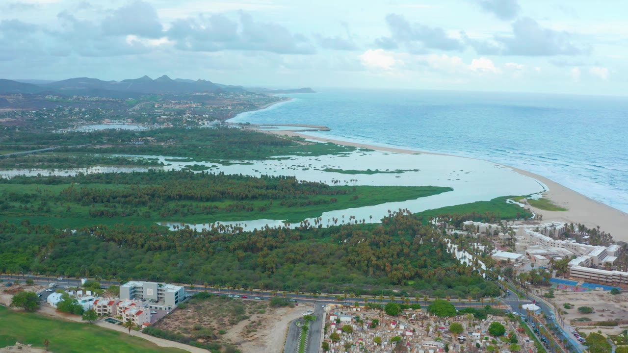 San Jose del Cabo aerial frame showing the ocean on the right, lagoon water and greenery across the center, roads and low buildings along the bottom edge, hills in the distance, and no visible people.